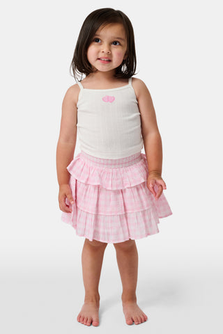 The Kids Hearts Pointelle Bridget Cami by Roller Rabbit is a white sleeveless camisole with thin straps and a small pink heart-shaped patch at the center of the chest. Crafted from soft pointelle cotton, it features a delicate scoopneck design. The background is light gray.