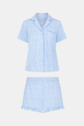 A woman stands barefoot in Roller Rabbit’s Lovely Monkey Wyatt Polo Pajamas—light blue Pima cotton with a button-up top and ruffled shorts, one hand in her pocket, against a plain white background.