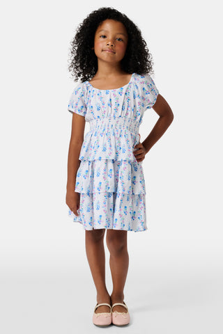 Four young girls stand and chat in front of white lockers, wearing pastel kids' dresses. One girl wears the Roller Rabbit Kids Naia Floral Stripe Alana Dress and holds a backpack and books, appearing to be in a school hallway. Another girl holds paper over her head.