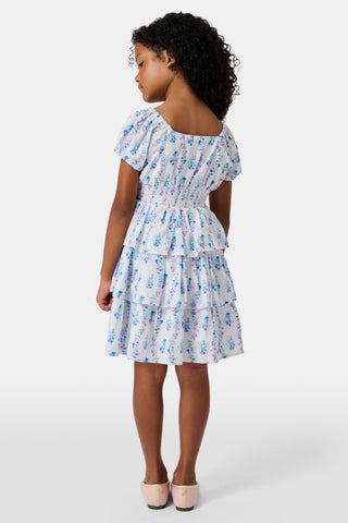 Four young girls stand and chat in front of white lockers, wearing pastel kids' dresses. One girl wears the Roller Rabbit Kids Naia Floral Stripe Alana Dress and holds a backpack and books, appearing to be in a school hallway. Another girl holds paper over her head.