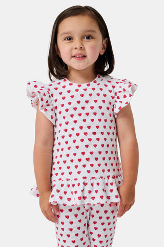 A child with long blonde hair wears the Roller Rabbit Kids Hearts Riley Set made from Pima Waffle fabric, featuring red hearts and ruffled cap sleeves, while holding a notebook with a decorative cover and standing in front of lockers.