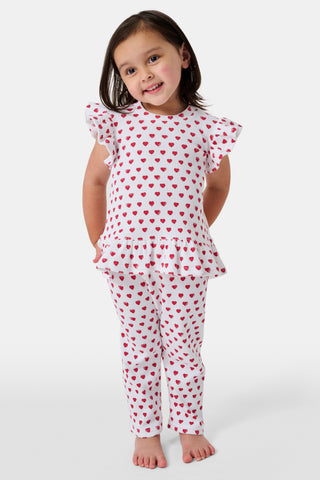 A child with long blonde hair wears the Roller Rabbit Kids Hearts Riley Set made from Pima Waffle fabric, featuring red hearts and ruffled cap sleeves, while holding a notebook with a decorative cover and standing in front of lockers.