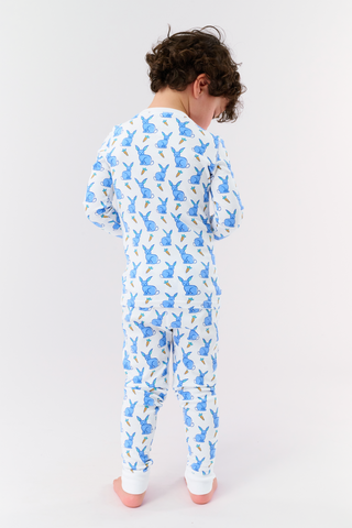 A young child with curly hair stands barefoot in white Kids Romy Rabbit Pajamas by Roller Rabbit, featuring a blue bunny print, looking at the camera and touching their mouth with one finger.