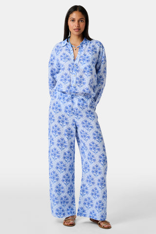 A woman stands against a plain light background wearing the Roller Rabbit Indigo Garden Boyfriend Shirt, a button-up long-sleeve pajama top in a blue floral print paired with matching wide-leg pants.