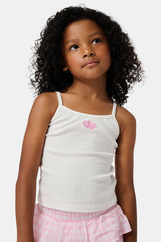 The Kids Hearts Pointelle Bridget Cami by Roller Rabbit is a white sleeveless camisole with thin straps and a small pink heart-shaped patch at the center of the chest. Crafted from soft pointelle cotton, it features a delicate scoopneck design. The background is light gray.