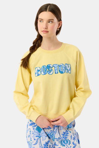 A woman with long brown hair in a braid wears the Roller Rabbit Boston Applique Crewneck and white and blue floral pants, standing against a plain light background.