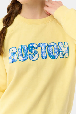 A woman with long brown hair in a braid wears the Roller Rabbit Boston Applique Crewneck and white and blue floral pants, standing against a plain light background.