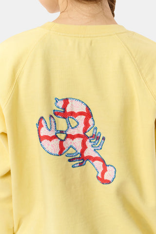 A woman with long brown hair in a braid wears the Roller Rabbit Boston Applique Crewneck and white and blue floral pants, standing against a plain light background.