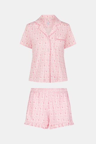 A woman with long curly hair wears Roller Rabbit Monkey Rosebud Wyatt Polo Pajamas—a light pink short-sleeve button-up shirt and shorts set with a small paisley pattern—standing against a plain light gray background.
