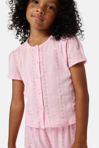 A young girl in a rose pink dress smiles while kneeling on the floor, holding a colorful folder. A pink backpack hangs nearby in a classroom or locker area, and she wears her cozy Roller Rabbit Kids Daisy Heart Stripe Diana Cardigan.