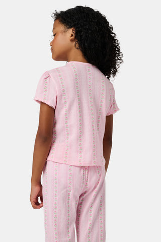 A young girl in a rose pink dress smiles while kneeling on the floor, holding a colorful folder. A pink backpack hangs nearby in a classroom or locker area, and she wears her cozy Roller Rabbit Kids Daisy Heart Stripe Diana Cardigan.