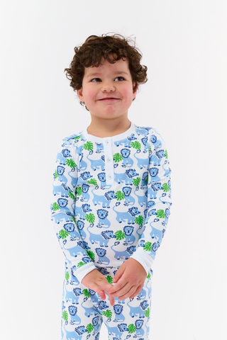 A young child with curly brown hair smiles while wearing Kids Leo The Lion Pajamas—soft Pima cotton PJs in blue and green, featuring playful lions and leaves—standing against a plain white background.