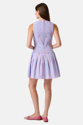 A young girl wearing the Roller Rabbit Kids Sweet Stripe Casey Dress stands in front of lockers, smiling and holding a folder decorated with colorful raindrops.
