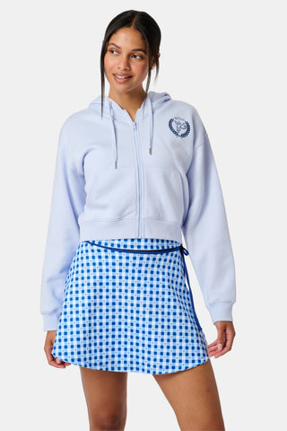 A woman wears the Roller Rabbit RR Athletic Club Cropped Zip Up—a pastel blue terry hoodie with an athletic club logo on the chest—paired with a blue-and-white checkered skirt, standing against a white background.
