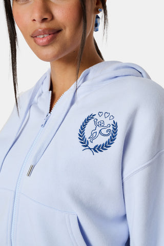 A woman wears the Roller Rabbit RR Athletic Club Cropped Zip Up—a pastel blue terry hoodie with an athletic club logo on the chest—paired with a blue-and-white checkered skirt, standing against a white background.
