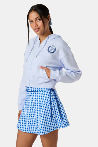 A woman wears the Roller Rabbit RR Athletic Club Cropped Zip Up—a pastel blue terry hoodie with an athletic club logo on the chest—paired with a blue-and-white checkered skirt, standing against a white background.