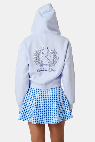 A woman wears the Roller Rabbit RR Athletic Club Cropped Zip Up—a pastel blue terry hoodie with an athletic club logo on the chest—paired with a blue-and-white checkered skirt, standing against a white background.
