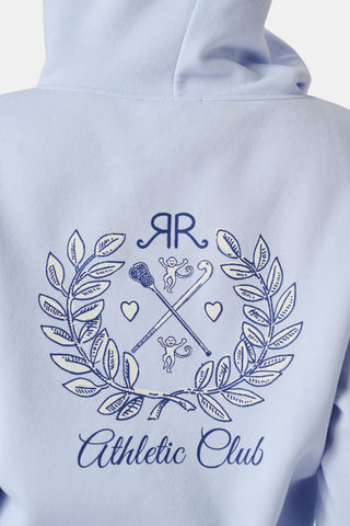 A woman wears the Roller Rabbit RR Athletic Club Cropped Zip Up—a pastel blue terry hoodie with an athletic club logo on the chest—paired with a blue-and-white checkered skirt, standing against a white background.