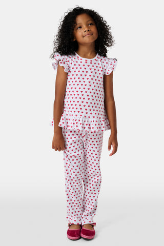 A child with long blonde hair wears the Roller Rabbit Kids Hearts Riley Set made from Pima Waffle fabric, featuring red hearts and ruffled cap sleeves, while holding a notebook with a decorative cover and standing in front of lockers.
