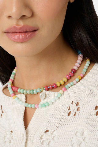 A woman wears Roller Rabbit’s Beaded Necklace Stack—one necklace with pastel beads and a white flower charm, the other with colorful beaded chains—paired with a white knit top featuring floral cutouts. Only her lower face and neck are visible.