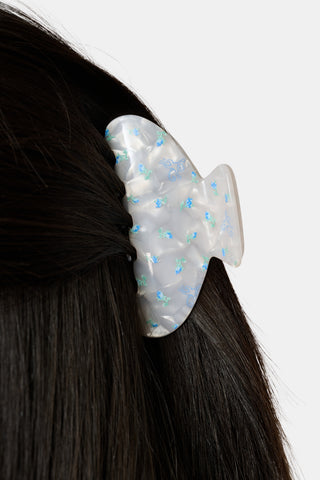 A close-up of dark hair secured with the Roller Rabbit Mini Monkey Blossom Claw Clip, a white, iridescent acrylic hair accessory adorned with small blue and green designs. The background is plain white.