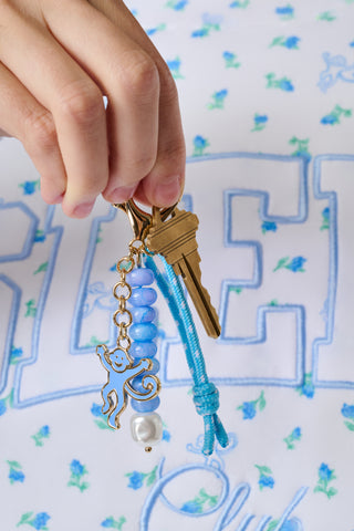 A hand holds two gold keys attached to the Roller Rabbit Monkey Beaded Keychain, featuring blue beads, a smiling monkey charm, a pearl bead, and a blue string, set against a white and blue floral patterned background.
