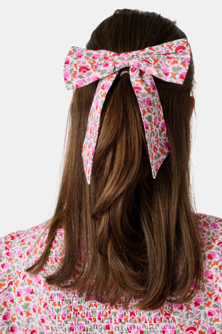 The Garden Gate Floral Hair Bow by Roller Rabbit is a large cotton bow with long tails, showcasing a vibrant pattern of pink, red, and orange flowers with green leaves on a white background.