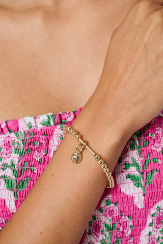 The Roller Rabbit Beaded Charm Bracelet features round, shiny gold beads evenly spaced in a circular design and joined by a sleek cylindrical clasp at the bottom. This elegant brass bracelet echoes the timeless style of classic foundation charm bracelets and is presented against a plain white background.