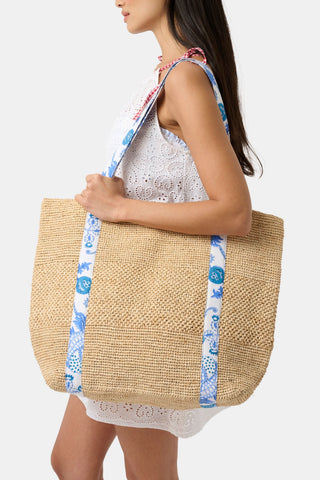 A woman in a sleeveless white eyelet dress carries the Amanda Woven Beach Bag by Roller Rabbit, featuring blue and white patterned shoulder straps. The large bag is shown from the side as she walks.