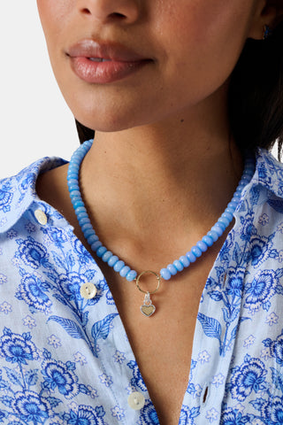 The Beaded Charm Necklace by Roller Rabbit features round, evenly spaced blue beads with a gold clasp, accented by a small gold ring pendant at the center. The necklace is shown against a plain white background.