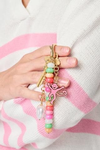 A hand holds gold keys on the Roller Rabbit Monkey Beaded Keychain, featuring colorful beads, a pink smiling monkey charm, and a white pearl charm. The person is wearing a white sweater with pink stripes.