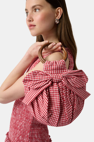 The Roller Rabbit Gingham Miranda Purse features red gingham fabric, a large red and white bow, gathered detailing, and a round bracelet handle, shown against a plain white background.
