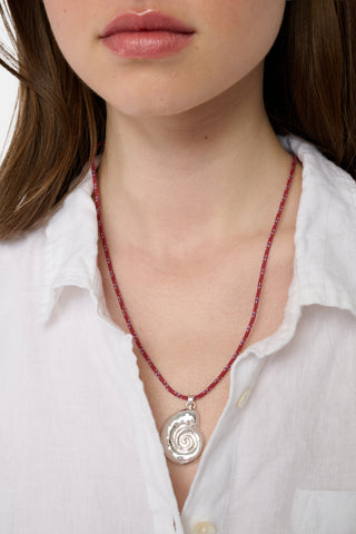 A woman, shown from the lower face down, wears a white button-up shirt and the Roller Rabbit Swirl Cord Necklace with a bold red cord and an eye-catching silver shell pendant.