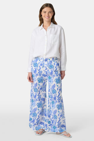Two women smile outdoors; one rides a bike in a blue and white shirt with white shorts, while the other walks beside her wearing Amanda Kaylee Pants. Green ivy covers the background fence as they enjoy a carefree day.