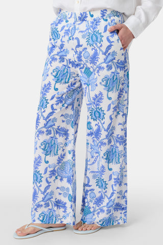 Two women smile outdoors; one rides a bike in a blue and white shirt with white shorts, while the other walks beside her wearing Amanda Kaylee Pants. Green ivy covers the background fence as they enjoy a carefree day.