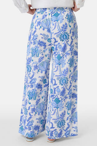 Two women smile outdoors; one rides a bike in a blue and white shirt with white shorts, while the other walks beside her wearing Amanda Kaylee Pants. Green ivy covers the background fence as they enjoy a carefree day.