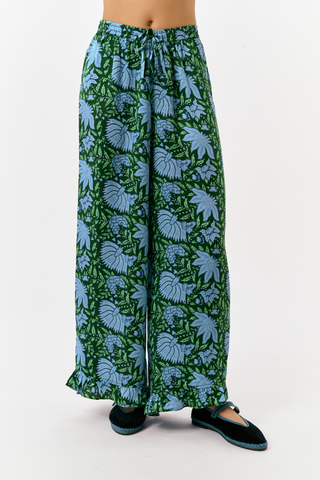 A woman wears the SAPPHIRE Antonia Floral Joanna Set, a washable silk two-piece featuring a green and blue floral print, a sleeveless ruffled-strap top, and wide-leg trousers.