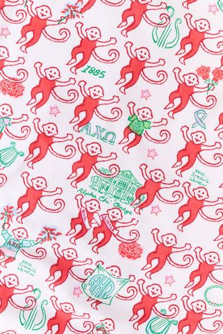 A woman stands outside with her arms crossed, wearing the Roller Rabbit Alpha Chi Omega Monkey Pajamas—a white Henley top and elastic waist pajama set featuring playful red and pink monkey patterns. She holds a matching quilted bag and carries a small heart-shaped blue and white polka dot purse.