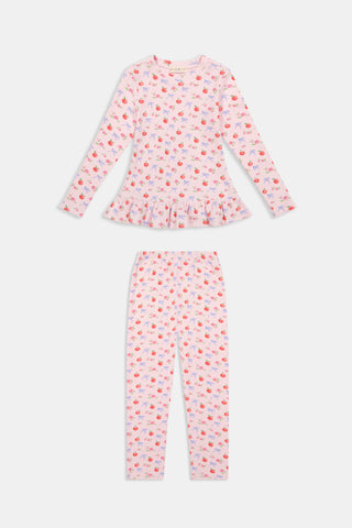 A young girl stands smiling, wearing the Roller Rabbit Kids Apple Bloom Elle Set—a pink girls’ loungewear outfit featuring an apple bloom print and a ruffled hem. She has long dark hair and is wearing light pink shoes, set against a plain white background.
