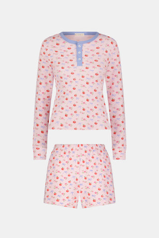 A woman wears the Roller Rabbit Apple Bloom Rosemary Set—a soft Pima cotton pajama ensemble featuring a pink long-sleeve top with a blue button placket and matching shorts, both adorned with a delicate red and pink apple bloom print. She stands with one hand in her pocket, gazing to the side.