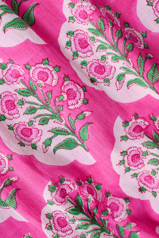 A person wearing the Roller Rabbit Ava Floral Bristol Dress—a pink cotton voile dress with a smocked bodice—has a green sweater draped over their shoulders and stands in a library near bookshelves.