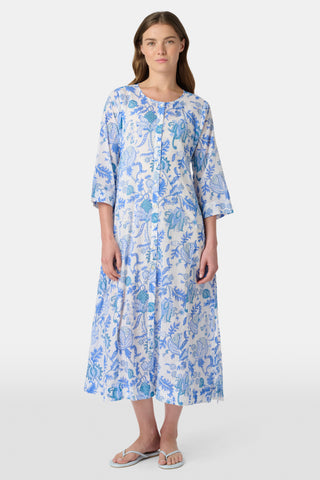 A woman stands in front of lush greenery, hands in her pockets and looking relaxed, wearing a straw hat and the Amanda Kurta—a blue-and-white floral piece made from 100% cotton.