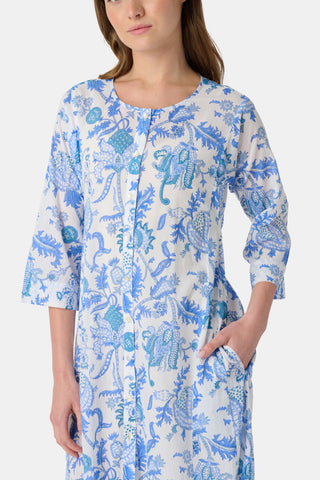 A woman stands in front of lush greenery, hands in her pockets and looking relaxed, wearing a straw hat and the Amanda Kurta—a blue-and-white floral piece made from 100% cotton.