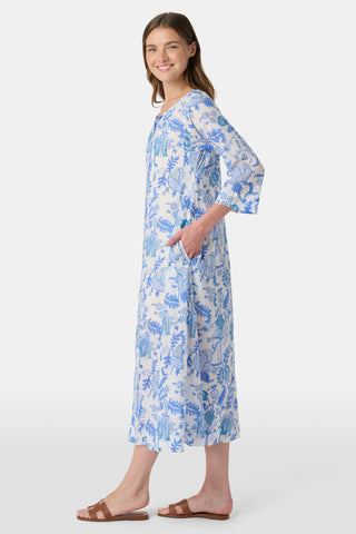 A woman stands in front of lush greenery, hands in her pockets and looking relaxed, wearing a straw hat and the Amanda Kurta—a blue-and-white floral piece made from 100% cotton.