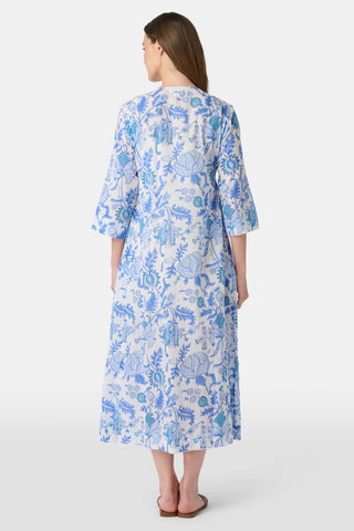A woman stands in front of lush greenery, hands in her pockets and looking relaxed, wearing a straw hat and the Amanda Kurta—a blue-and-white floral piece made from 100% cotton.