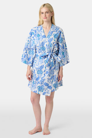 A woman stands in front of lush greenery, hands in her pockets and looking relaxed, wearing a straw hat and the Amanda Kurta—a blue-and-white floral piece made from 100% cotton.