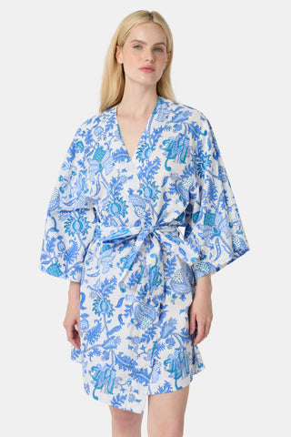 A woman stands in front of lush greenery, hands in her pockets and looking relaxed, wearing a straw hat and the Amanda Kurta—a blue-and-white floral piece made from 100% cotton.