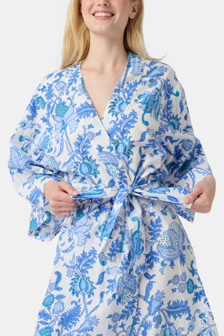 A woman stands in front of lush greenery, hands in her pockets and looking relaxed, wearing a straw hat and the Amanda Kurta—a blue-and-white floral piece made from 100% cotton.