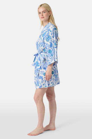 A woman stands in front of lush greenery, hands in her pockets and looking relaxed, wearing a straw hat and the Amanda Kurta—a blue-and-white floral piece made from 100% cotton.