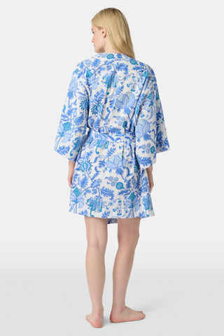 A woman stands in front of lush greenery, hands in her pockets and looking relaxed, wearing a straw hat and the Amanda Kurta—a blue-and-white floral piece made from 100% cotton.